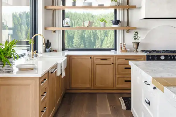 Modern kitchen with wooden cabinets, white countertops, and a farmhouse sink. Open shelves hold plants and kitchenware, while a window offers a view of trees. For peace of mind, keep "plumber repair" services handy to ensure everything runs smoothly in your stylish space.