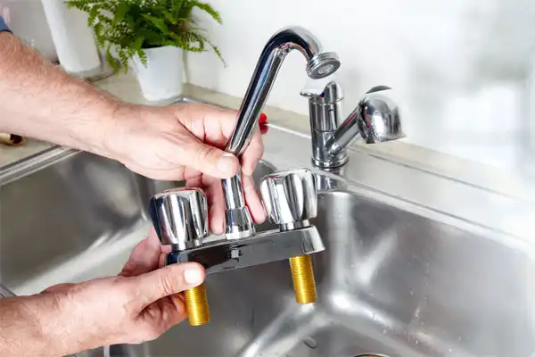 A person holds a detached two-handle faucet over a kitchen sink, preparing for installation. A plant and another faucet are visible in the background.