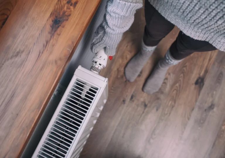 Person wearing gray sweater and gloves adjusts a radiator valve in a room with wooden flooring.