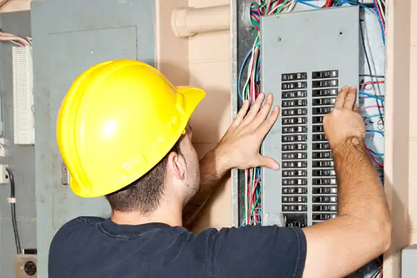 Electrician wearing a yellow hard hat inspects a breaker panel with exposed wires, using both hands to adjust switches.