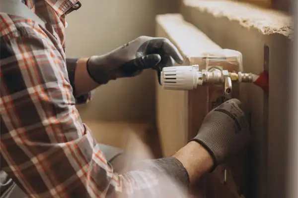 Person in gloves adjusting a valve on a radiator, wearing a plaid shirt.