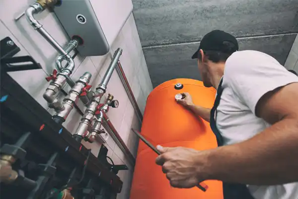 A technician wearing a cap works on an orange industrial tank, adjusting a valve with a wrench in a utility room with visible pipes above.