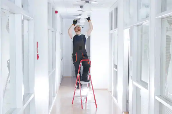 Person standing on a ladder fixing a ceiling light in a bright hallway.