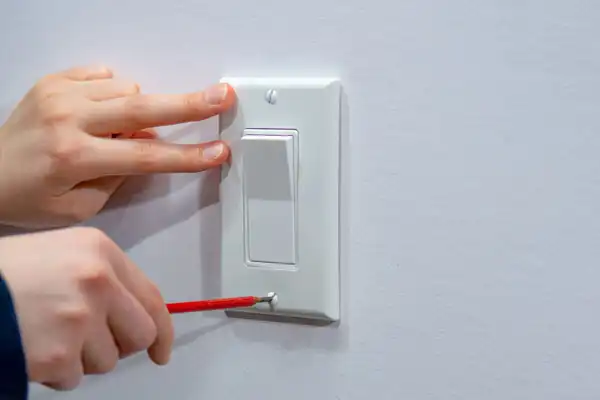 Hand holding a screwdriver, adjusting a white light switch on a wall, while another hand steadies the switch cover.