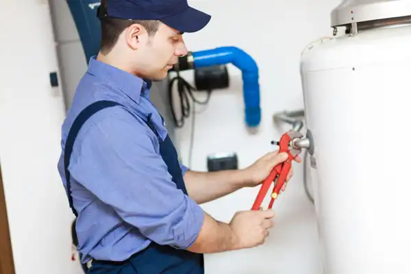 A person in a blue uniform and cap uses a wrench to adjust a valve on a large white tank in a utility area.