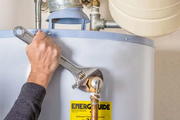 A person tightens a connection on a water heater using a large adjustable wrench. The heater has an "EnerGuide" label attached.