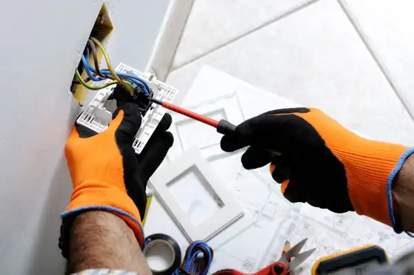 Person wearing orange gloves using a screwdriver to repair a wall electrical outlet, surrounded by tools and wires.